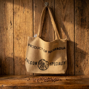 A natural jute tote bag with black printed text reading 'PRODUCT OF NICARAGUA' and 'FALCON SPECIALTY' hanging against a white paneled wall.