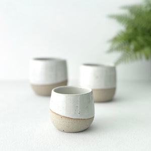 Three ceramic cups with a natural finish on a light surface with a blurred plant in the background